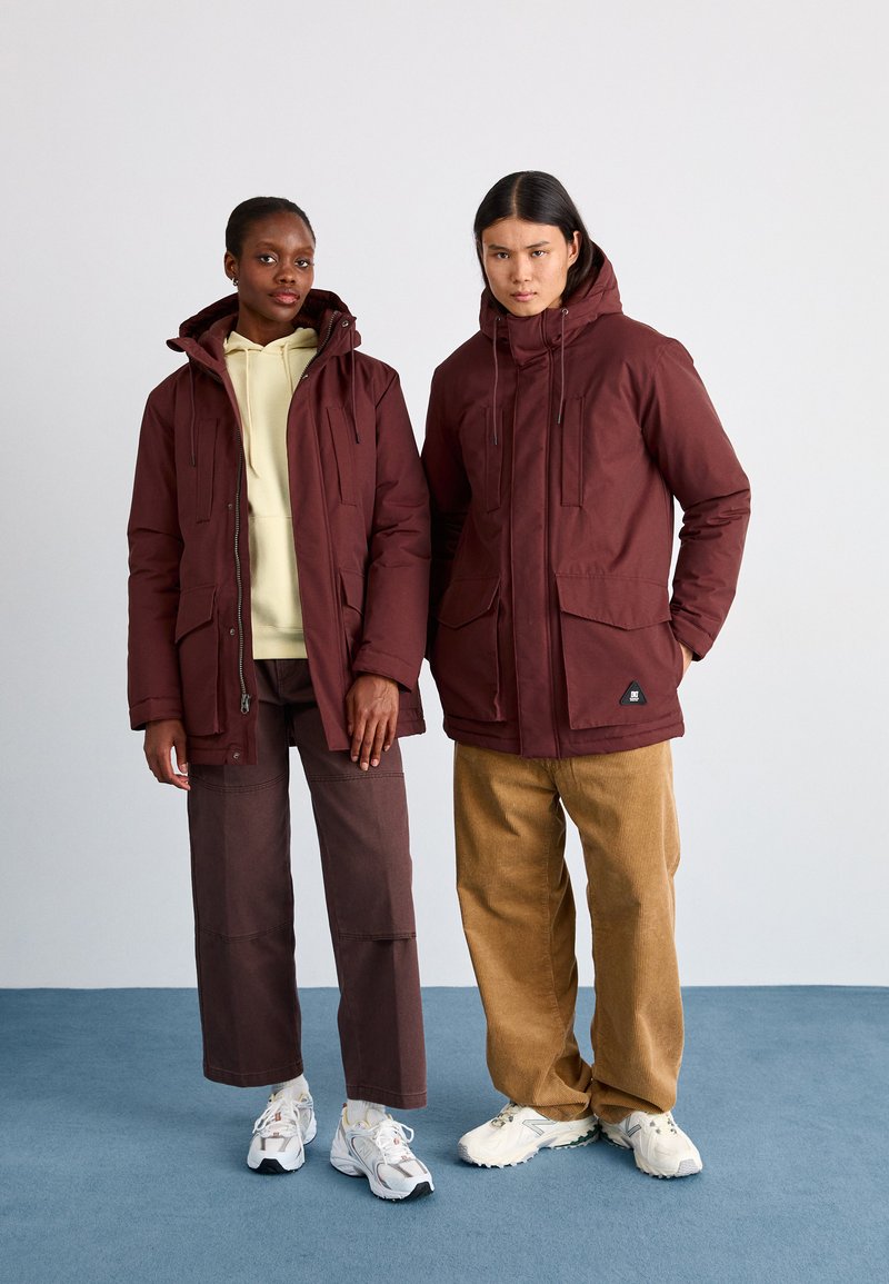 Chaquetas acolchadas burdeos con capuchas, bolsillos frontales y cremalleras. El modelo masculino lleva pantalones de pana beige, mientras que el modelo femenino lleva pantalones marrones.