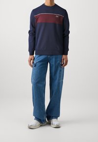 Felpa blu navy con pannello di petto bordeaux, piping bianco e logo. Abbinata a jeans cargo a gamba larga in denim blu e sneakers argentate.