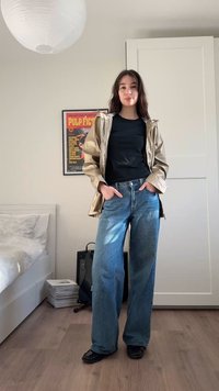 Jeune femme aux longs cheveux portant une veste beige, un chemisier noir et un jean bleu, debout dans une chambre avec une affiche de "Pulp Fiction" au mur.