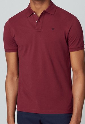 Mann trägt ein burgunderfarbenes Poloshirt mit kurzen Ärmeln, zwei geöffneten Knöpfen und einem kleinen gestickten Logo auf der linken Brust.