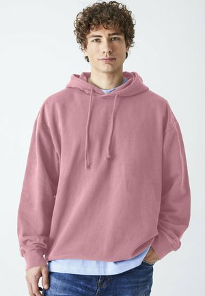 Rosa Kapuzenpullover mit Fronttasche, Kordelzugkapuze und lockerer Passform. Über einem hellblauen Shirt getragen und mit blauen Jeans kombiniert.