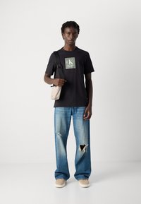 Joven con una camiseta negra de Calvin Klein, jeans rotos azules, zapatillas beige y un bolso cruzado beige, frente a un fondo sencillo.