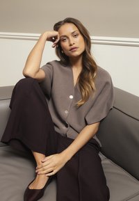 Brun stickad kofta med korta ärmar, med tre metallknappar. Matchad med mörkbruna vida byxor och burgunderröda ballerinaskor på en grå soffa.