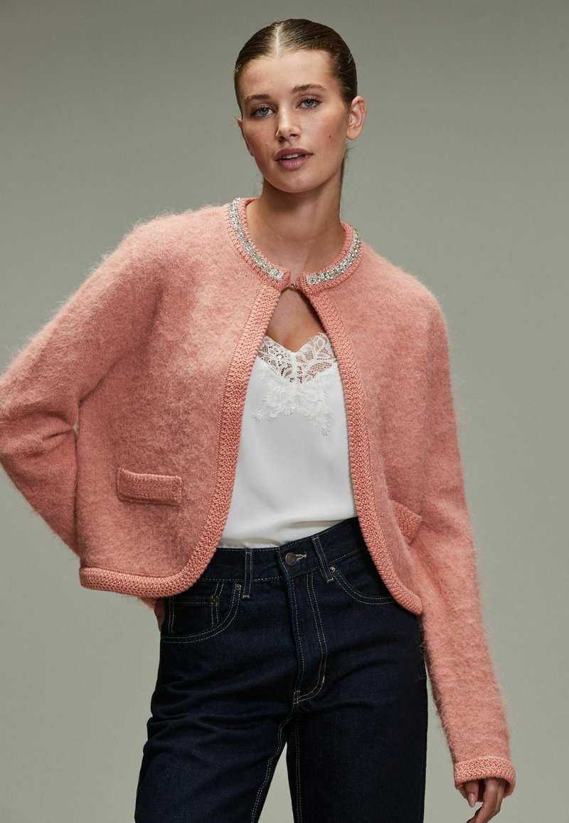 Light pink cropped cardigan made of fluffy material, featuring a knitted trim, round neckline, and a single front pocket with a subtle sheen.
