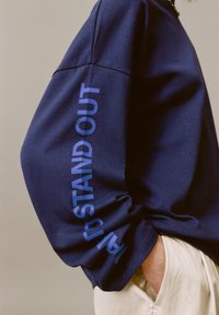Felpa blu navy a maniche lunghe con la scritta "STAND OUT" in chiaro azzurro in grassetto sul polsino. Tessuto liscio con vestibilità comoda.