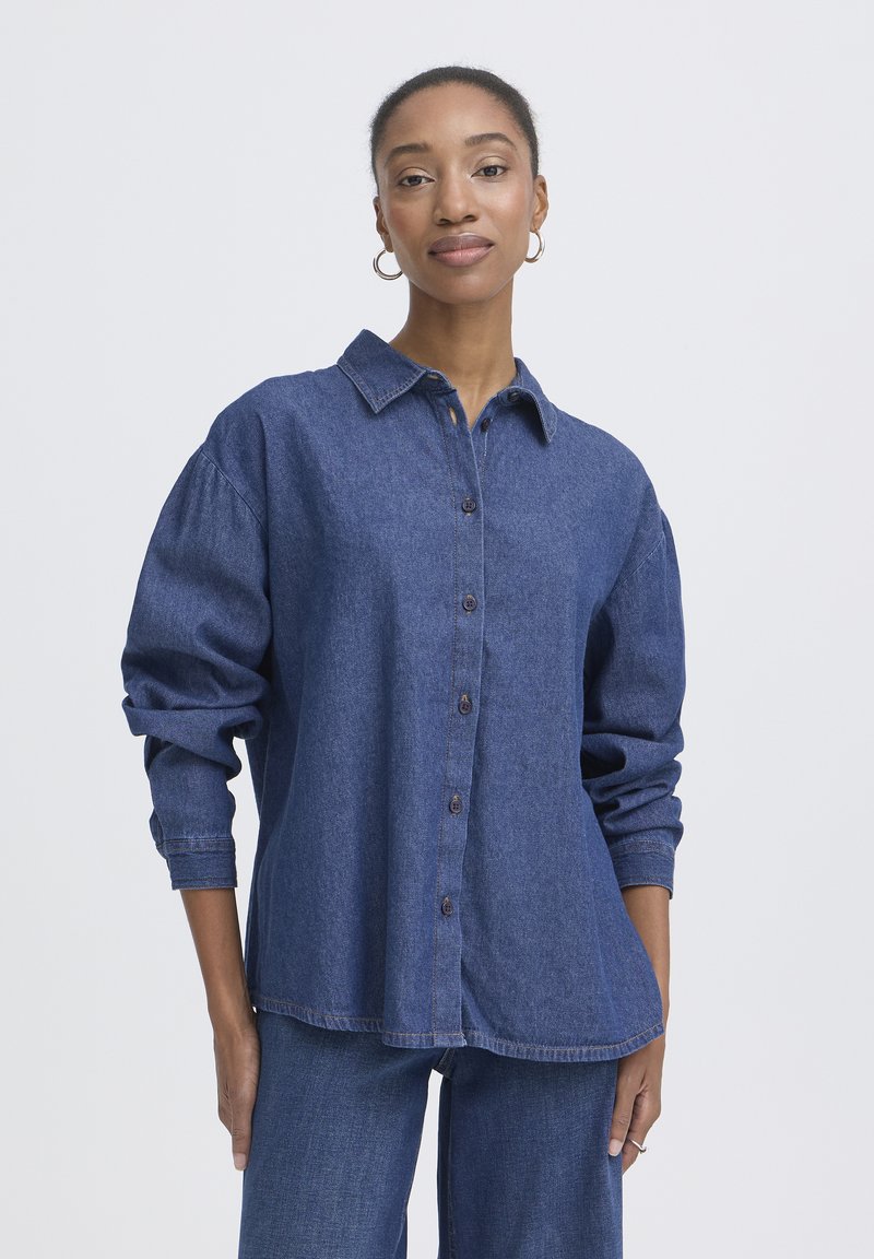 Chemise en denim bleu foncé, avec fermeture boutonnée sur le devant, col chemise, et manches amples aux poignets retroussés. Texture lisse.