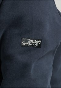 Navy sweatshirt fabric with a woven black label featuring white text: "Styled & Made by Superdry Manufacturing Co."