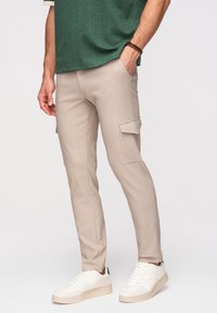 Homme portant un pantalon cargo beige avec des poches latérales, des baskets blanches et une chemise verte à manches courtes, debout avec une main dans la poche.