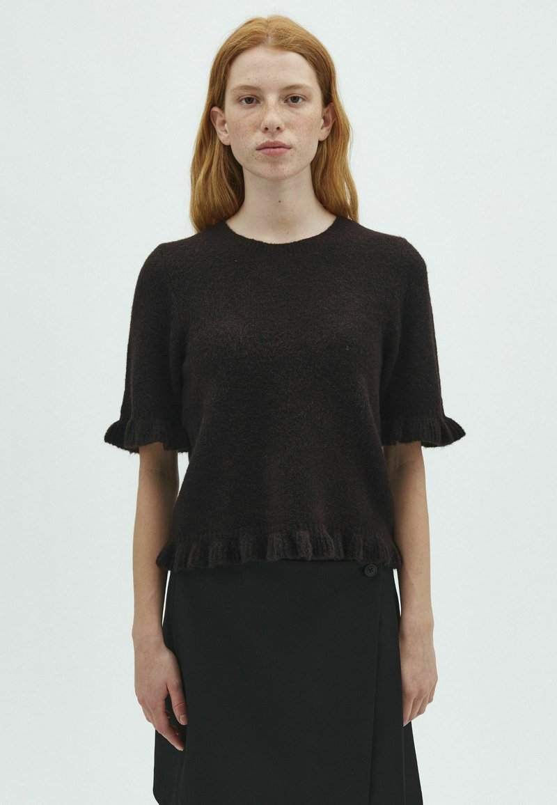 Jeune femme aux cheveux roux portant un pull noir à manches courtes avec des bords volantés et une jupe noire, debout devant un fond blanc uni.