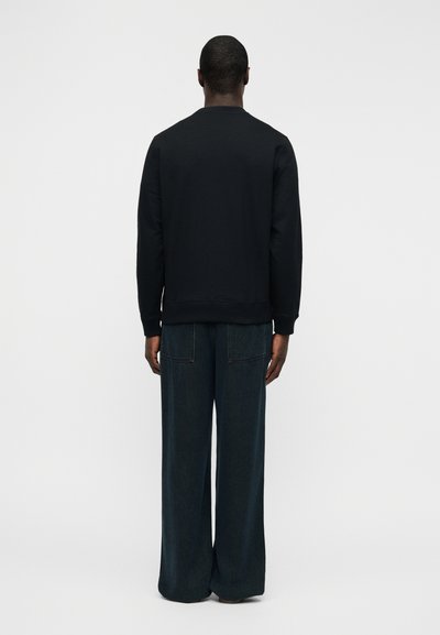 Sweat-shirt noir avec un col rond et des poignets côtelés, porté avec un pantalon en denim sombre à coupe ample. Vue de dos unie, sans motifs visibles.