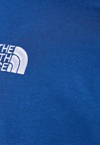 Modrá bavlněná tkanina s bílým vyšívaným logem "The North Face" s zakřiveným designem. Hladká textura, detailní pohled.