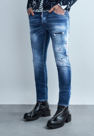 Στενά φθαρμένα μπλε τζιν φορεμένα με μαύρα δερμάτινα μποτάκια αστραγάλου με φερμουάρ μπροστά και τακούνια τύπου block, με τα χέρια στις τσέπες.