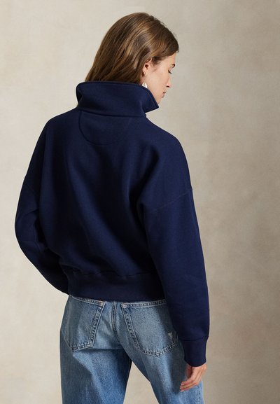 Mujer de cabello castaño claro con una sudadera azul marino de cuello alto y jeans azules, de perfil frente a un fondo liso.