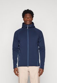 Tumesinine tõmblukuga jakk kapuutsiga, valmistatud venivast kangast, millel on kontrastsed helesinised õmblused, kaks küljetaskut ja vormitud disain.