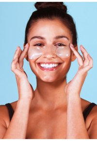 Hydrogel-Augenkissen, die unter den Augen einer lächelnden Frau mit glänzender Oberfläche auf einem Hautton aufgetragen sind, vor einem hellblauen Hintergrund.