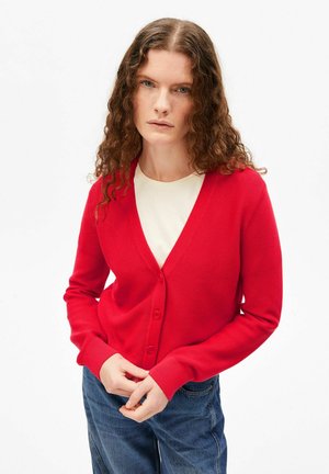 Vrouw met krullend haar, gekleed in een rode cardigan met knopen over een wit shirt en blauwe jeans, staand met gevouwen handen voor zich tegen een witte achtergrond.