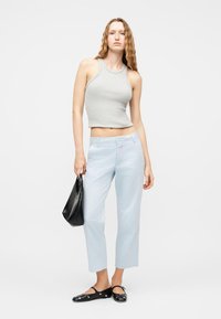 Γυναίκα με κυματιστά μαλλιά που φοράει γκρι αμάνικο τοπ, ανοιχτό μπλε κοντά παντελόνια, μαύρες μπαλαρίνες με τρουκς, κρατώντας μαύρη τσάντα χειρός.