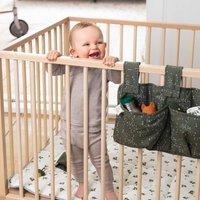 Holz-Kinderbett mit vertikalen Stäben, einer weichen weißen Matratze mit grünem Blumenmuster und einer grünen Aufbewahrungstasche, die Spielzeug und eine Flasche hält.