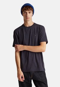 Mladý muž s modrou čepicí a tmavě šedým tričkem, stojící se zkříženou jednou rukou přes druhou na jednobarevném pozadí.