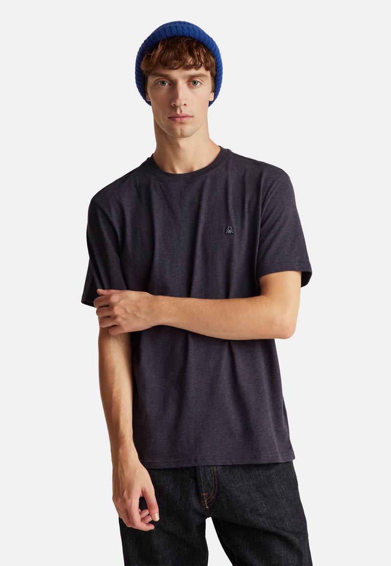 Mladý muž s modrou čepicí a tmavě šedým tričkem, stojící se zkříženou jednou rukou přes druhou na jednobarevném pozadí.