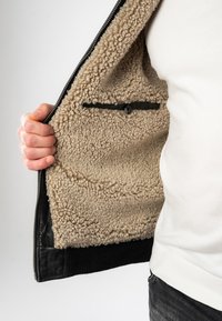 Schwarze Zip-Jacke mit beigem, plüschigem Innenfutter, ausgestattet mit einer versteckten Tasche mit Knopfverschluss. Abgerundet mit gerippten schwarzen Bündchen.