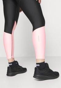 Černé a růžové sportovní legíny s přiléhavým střihem, které mají růžovou část na spodní noze a nenápadný branding v blízkosti kotníku.
