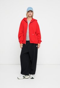 Felpa rossa con zip, t-shirt grigia, pantaloni neri a gamba larga, sneakers bianche con dettagli verdi e un cappellino azzurro chiaro. Branding minimale visibile.