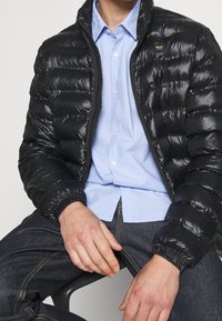 Giacca a vento nera con esterno lucido e trapuntato, colletto rialzato, chiusura con zip e polsi elasticizzati, indossata sopra una camicia azzurro chiaro con bottoni.