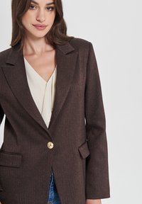 Blazer marrone con texture, un singolo bottone dorato, revers a scollo e tasche frontali, indossato sopra una blusa di colore crema chiaro.