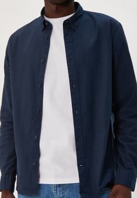 Tengerész kék gombos ing gallérral, puha anyagból készült, laza szabású, és alatta egy fehér környakú pólóval van styled.