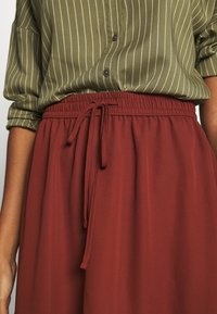 Camisa de botones a rayas verdes con mangas enrolladas, combinada con pantalones rojos holgados que tienen una cintura elástica y cordón.