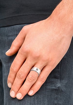 Un primo piano di una mano appoggiata su jeans scuri, che mostra un anello d'argento con una fascia decorata e una piccola decorazione sulla parte superiore.