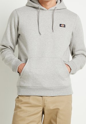 Muž v světle šedé mikině s kapucí, kangaroo kapsou a logem na hrudi, v kombinaci s béžovými kalhotami.