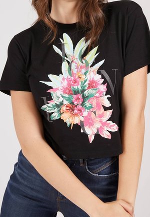 Woman wearing black cropped t-shirt with colorful floral print and faded text, paired with blue jeans, posing with arms crossed.