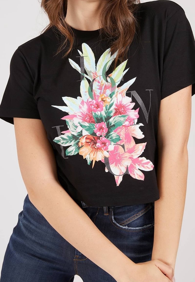 Femme portant un t-shirt court noir avec un imprimé floral coloré et un texte délavé, associé à un jean bleu, posant avec les bras croisés.