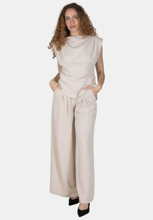 Donna con i capelli ricci che indossa una canotta beige e pantaloni beige a gamba larga, in piedi con le mani in tasca e scarpe nere a punta.