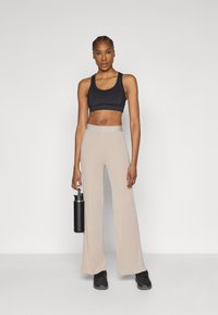 Musta urheilurintaliivi yhdistettynä beigen värisiin leveälahkeisiin housuihin, joissa on sileä pinta. Malli pitelee toisessa kädessään mustaa vesipulloa.