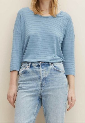 Mujer vistiendo una camiseta de rayas azul claro de manga larga metida en unos vaqueros de tiro alto de lavado claro, de pie contra un fondo neutro.