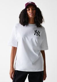 Camiseta de algodón blanca con corte oversize, mangas cortas y un logotipo negro de NY en el pecho. Llevada con una gorra color burdeos que presenta el mismo logotipo.