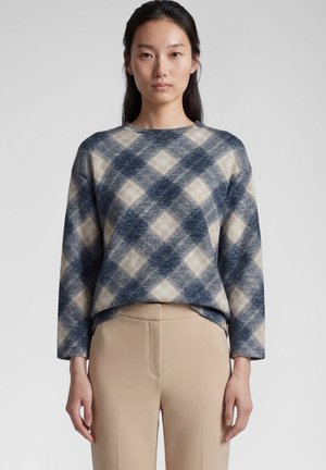 Young woman wearing a blue and beige plaid long-sleeve top and beige pants standing against a plain light background.