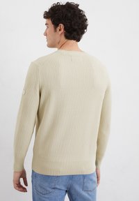 Beige gerippter Strickpullover mit langen Ärmeln, Rundhalsausschnitt und engem Saum. Das Modell trägt ihn mit hellblauen Jeans.