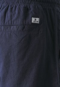 Detaily tmavě modré látky s zadní kapsou a bílou značkovou etiketou Jack & Jones blízko elastického pásku.