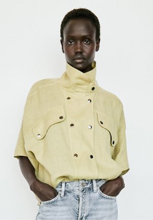 Dunkelhäutige Person trägt eine beige Jacke mit hohem Kragen und kurzen Ärmeln mit Knöpfen sowie hellblaue Jeans, Hände in den Taschen, neutraler Hintergrund.