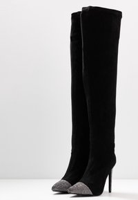 Mustat samettiset reisi-high saappaat, joissa on terävät varpaat, koristeelliset rhinestone-yksityiskohdat varpaan päällä ja korkea stiletto-korko.