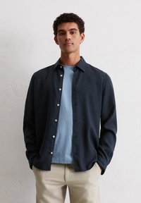Dunkelblaues Oberhemd mit entspannter Passform und langen Ärmeln, über einem hellblauen Rundhals-T-Shirt getragen, dazu beigen Hosen.