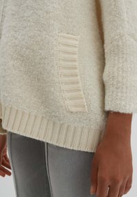 Kermanvalkoinen neulepaita, jossa on teksturoitu vartalo, resorireuna ja sivutasku vaakasuorin resoriraidoituksin.