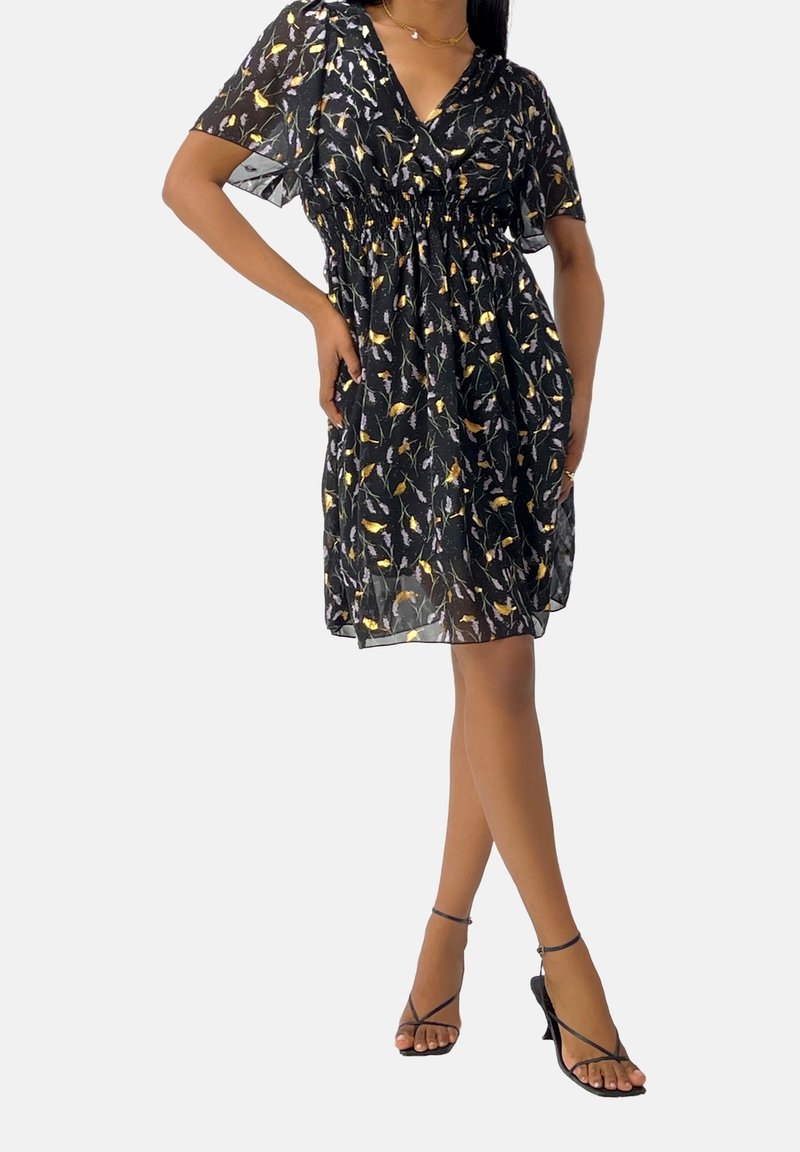 Femme portant une robe noire fleurie longueur genou et des sandales noires à talons avec lanières, debout avec une main sur la hanche.