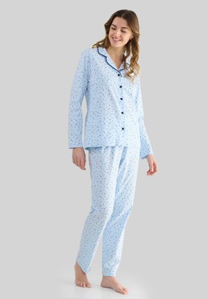 Mujer de pie descalza, vistiendo un pijama de botones de manga larga en color azul claro con un pequeño estampado floral oscuro y ribete oscuro en el cuello.