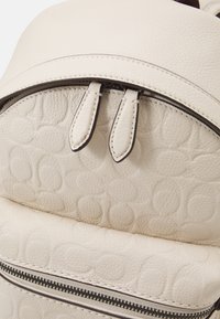 Beżowy skórzany plecak z tłoczonym wzorem, wyposażony w zamek błyskawiczny i dwa skórzane uchwyty. Gładka faktura z kontrastującym przeszyciem.