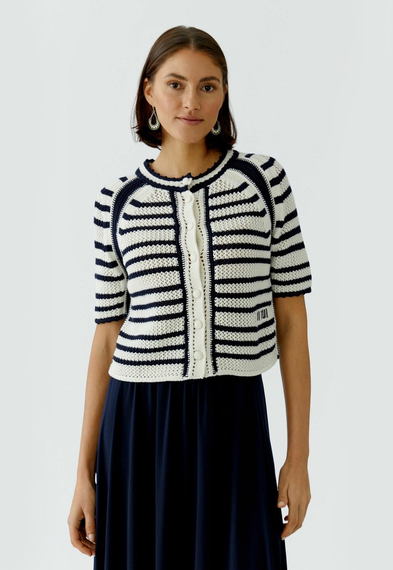 Femme portant un cardigan en tricot à manches courtes blanc à rayures bleu marine avec des boutons, associé à une jupe foncée et des boucles d'oreilles argentées.
